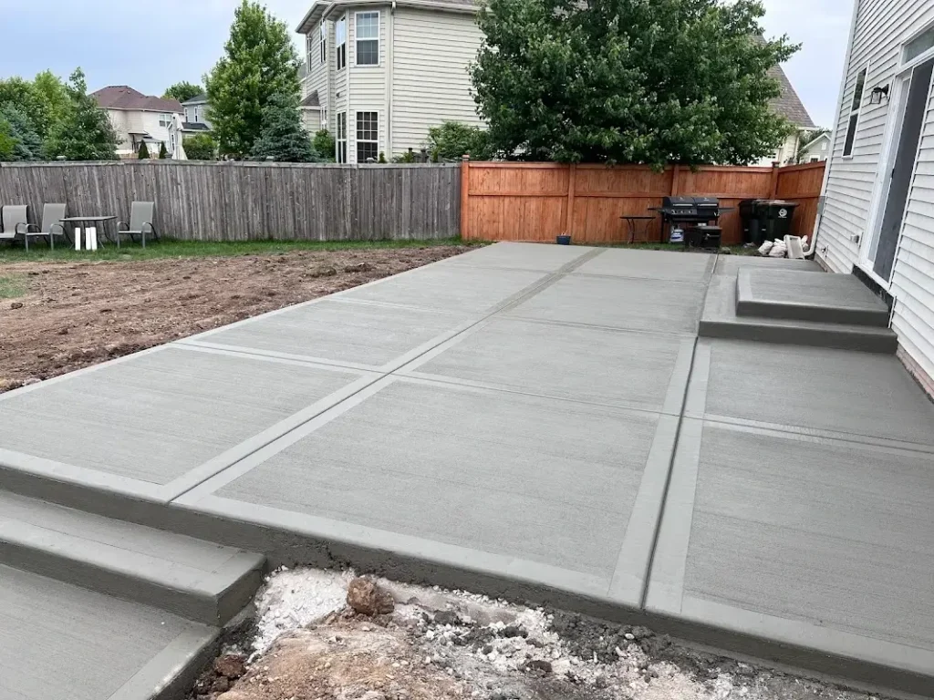 Newly poured concrete patio with clean expansion joints, featuring a smooth gray finish and steps, bordered by bare soil and wooden fencing in a residential backyard